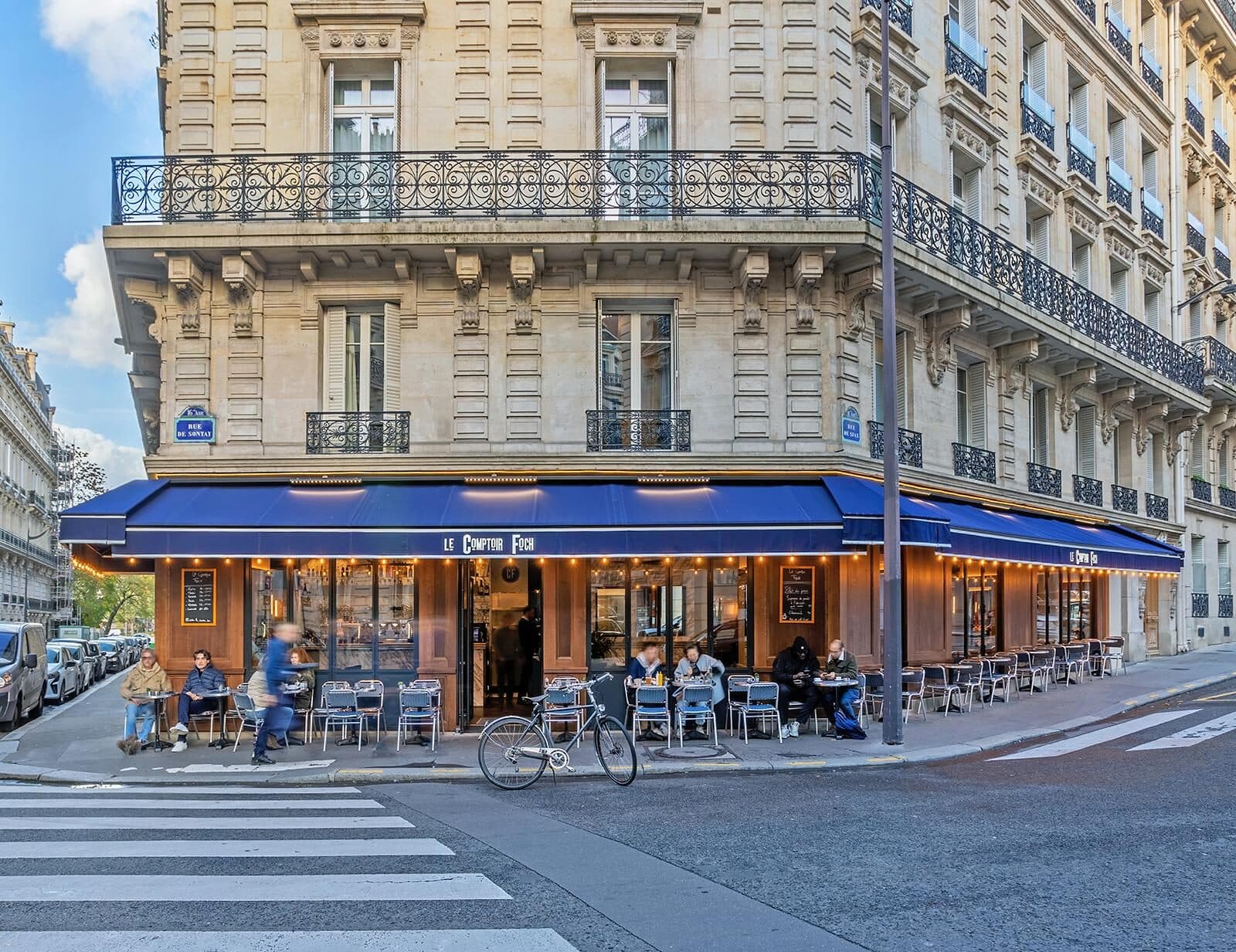 Façade en angle du Comptoir Foch, marquise bleu marine et terrasse animée dans un immeuble haussmannien du 16e arrondissement