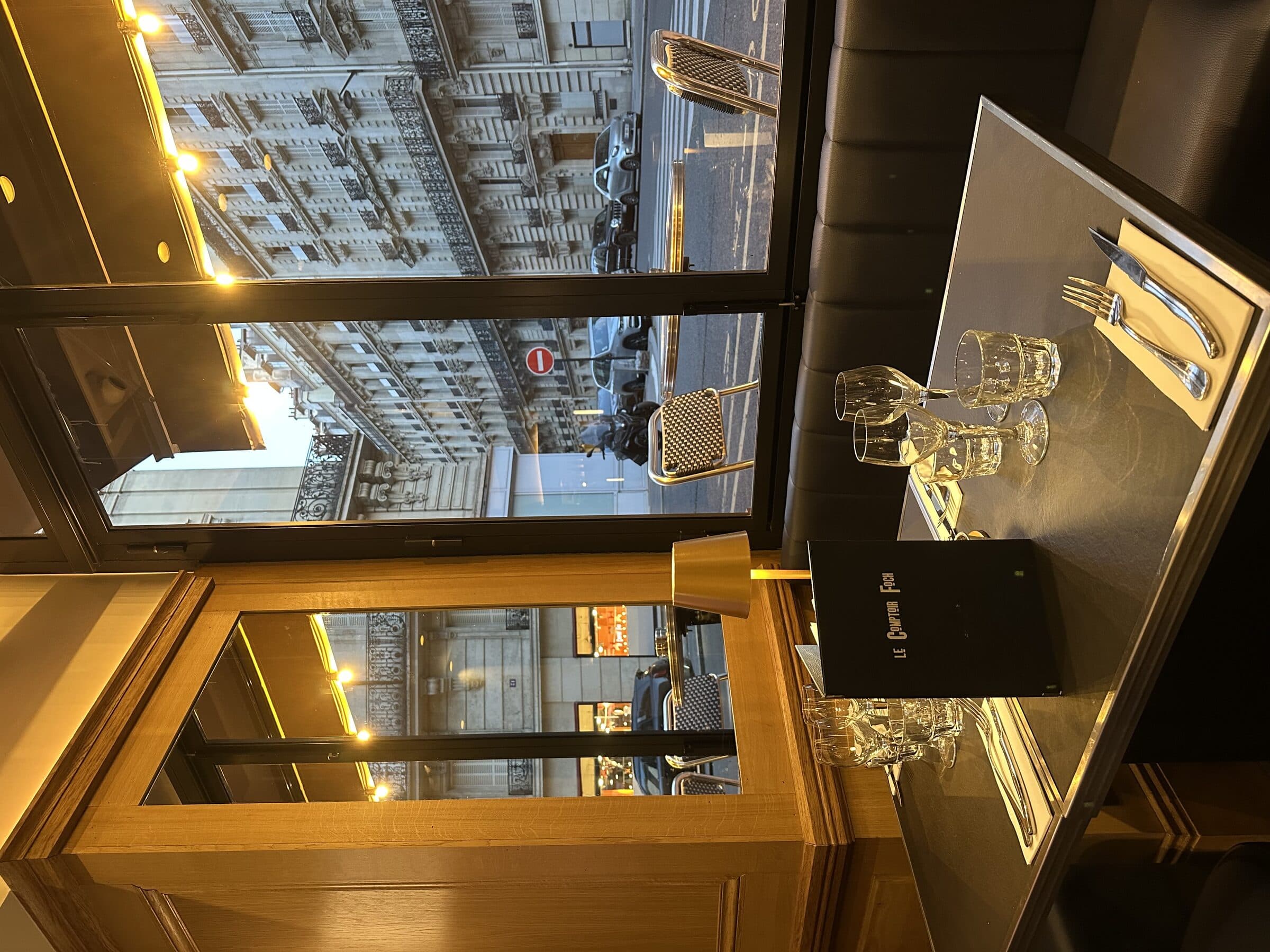 Vue intérieure du Comptoir Foch depuis le bar vers la baie vitrée, lumière ambrée et verres alignés au crépuscule