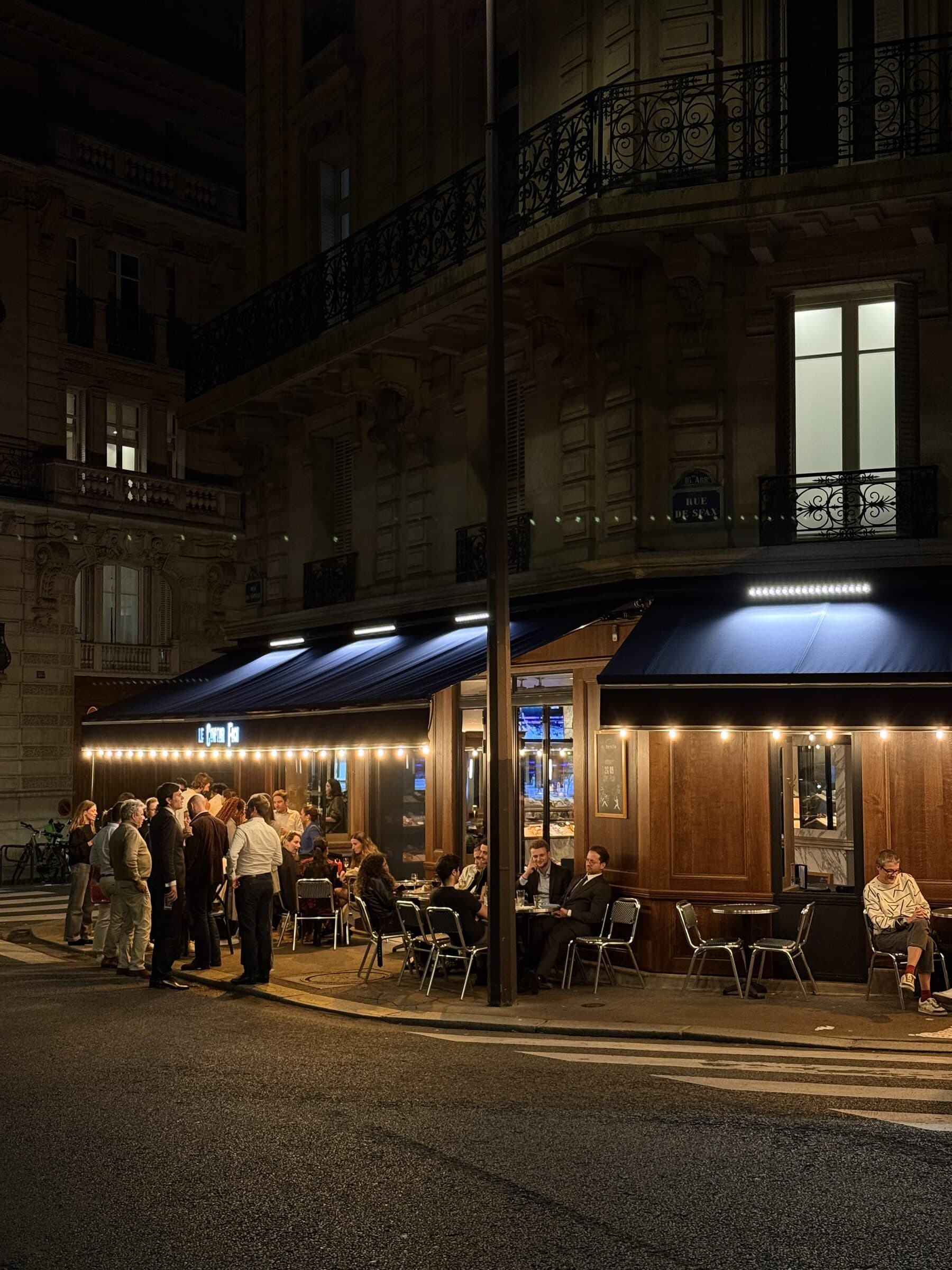 Terrasse nocturne animée du Comptoir Foch, guirlandes lumineuses, marquise bleu marine et clients en soirée parisienne